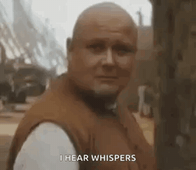 A Bald Man In A Brown Vest Is Standing In Front Of A Tree And Says `` I Hear Whispers '' . GIF