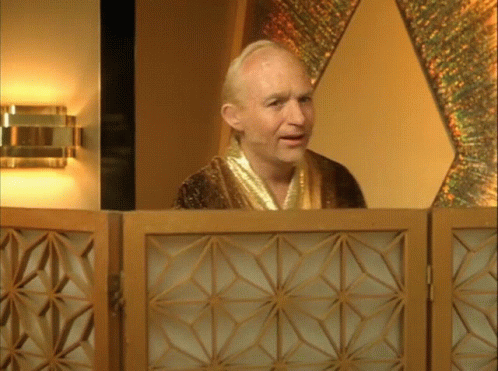 A Bald Man In A Gold Robe Stands Behind A Wooden Screen GIF