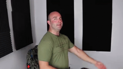 A Bald Man In A Green T-Shirt Is Standing In Front Of A Wall With Foam Panels . GIF