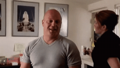 A Bald Man In A Grey Shirt Is Smiling While A Woman Stands Behind Him GIF