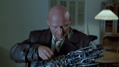 A Bald Man In A Leather Jacket And Tie Is Holding A Mechanical Arm GIF
