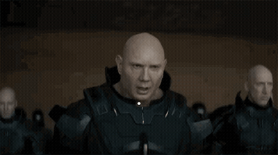 A Bald Man In A Military Uniform Is Standing In Front Of A Group Of Bald Men In A Dark Room . GIF
