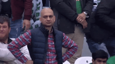 A Bald Man In A Plaid Shirt And Vest Is Sitting In A Crowd With His Hands On His Hips . GIF