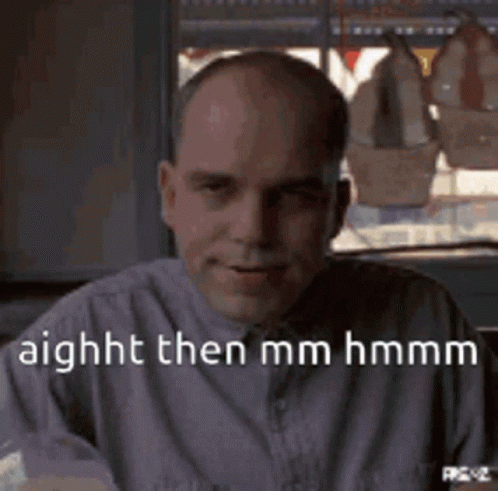 A Bald Man In A Purple Shirt Is Sitting In Front Of A Window And Smiling . GIF