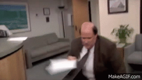 A Bald Man In A Suit And Tie Is Sitting At A Table In A Waiting Room .. GIF