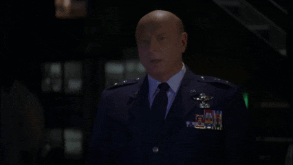 A Bald Man In A Suit And Tie Is Standing In Front Of A Sign That Says Godspeed Colonel GIF