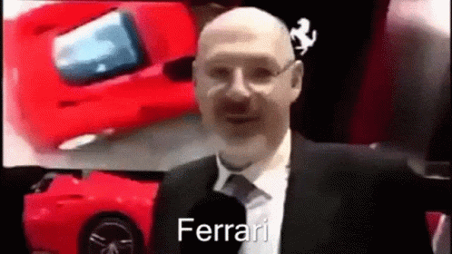 A Bald Man In A Suit And Tie Is Talking Into A Microphone While Standing In Front Of A Red Ferrari . GIF