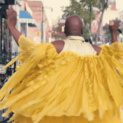 A Bald Man In A Yellow Dress Is Walking Down A Street GIF