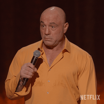 A Bald Man In An Orange Shirt Is Holding A Microphone In Front Of A Netflix Sign GIF