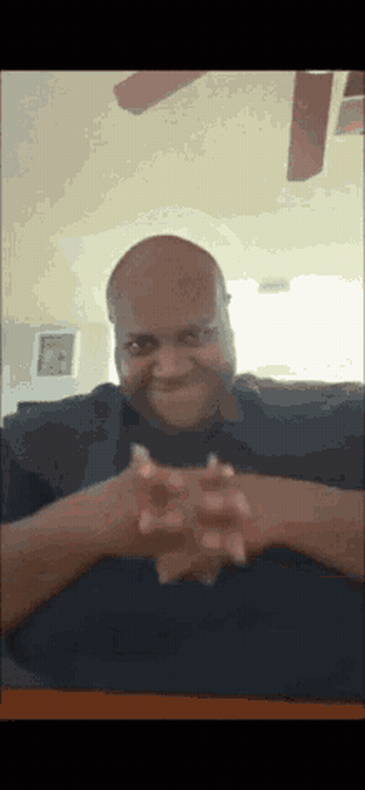 A Bald Man Is Sitting At A Table With His Hands Folded GIF