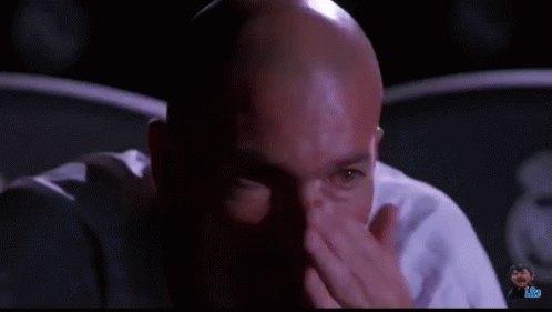 A Bald Man Is Sitting In A Dark Room And Looking At The Camera GIF