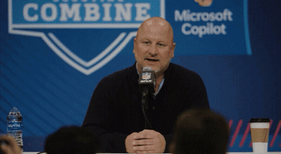A Bald Man Is Speaking Into A Microphone In Front Of A Sign That Says Combine GIF