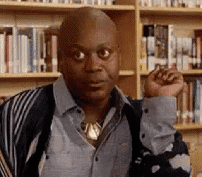 A Bald Man Is Standing In Front Of A Bookshelf In A Library With His Fist In The Air . GIF