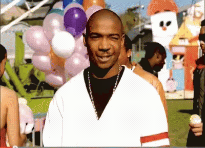 A Bald Man Stands In Front Of A Bunch Of Balloons At A Carnival GIF