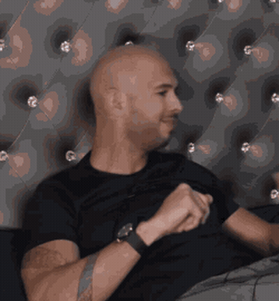 A Bald Man Wearing A Black Shirt And A Watch Is Sitting On A Couch GIF