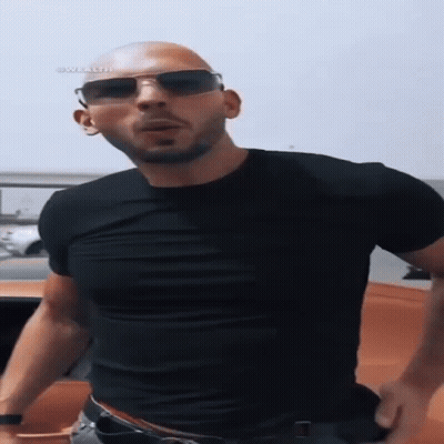 A Bald Man Wearing A Black Shirt And Sunglasses Is Standing In Front Of A Car . GIF