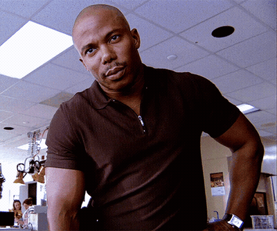 A Bald Man Wearing A Brown Polo Shirt And A Watch Stands In A Room GIF