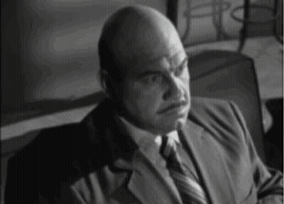 A Bald Man Wearing A Suit And Tie Is Sitting In A Chair GIF