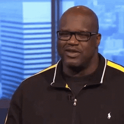 A Bald Man Wearing Glasses And A Black And Yellow Polo Shirt Is Standing In Front Of A Window . GIF