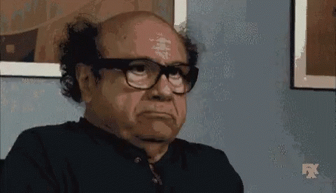 A Bald Man Wearing Glasses And A Black Shirt Is Sitting In Front Of A Wall . GIF