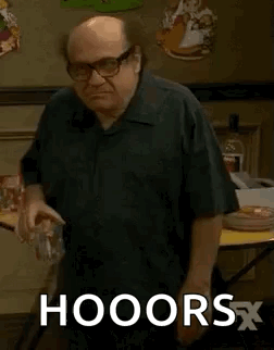 A Bald Man Wearing Glasses And A Black Shirt Is Standing In Front Of A Table And Holding A Glass Of Water . GIF