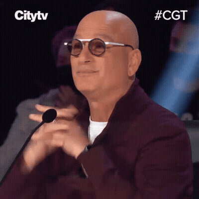A Bald Man Wearing Glasses And A Burgundy Jacket Is Clapping His Hands In Front Of A Microphone . GIF