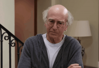 A Bald Man Wearing Glasses And A Gray Sweater Looks At The Camera GIF