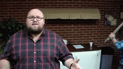 A Bald Man Wearing Glasses And A Plaid Shirt Stands In Front Of A Brick Wall GIF