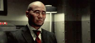 A Bald Man Wearing Glasses And A Suit Stands In A Dark Room GIF