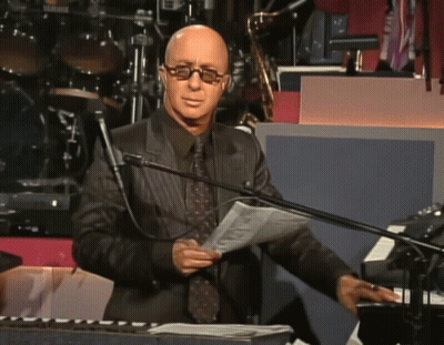 A Bald Man Wearing Glasses Stands In Front Of A Microphone Holding A Piece Of Paper GIF