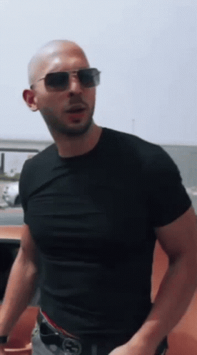 A Bald Man Wearing Sunglasses And A Black T-Shirt Is Standing In Front Of A Car . GIF