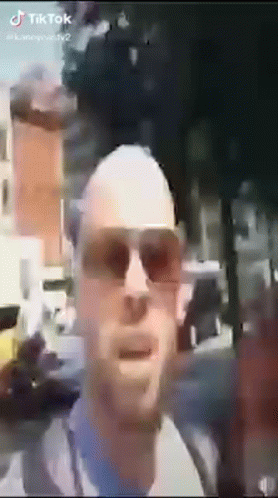A Bald Man Wearing Sunglasses Is Standing In Front Of A Building . GIF