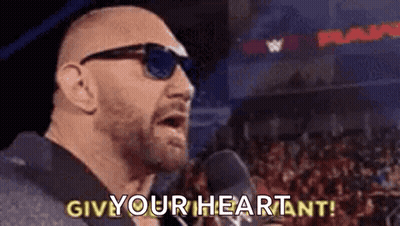 A Bald Man Wearing Sunglasses Is Standing In Front Of A Microphone And Saying `` Give Your Heart ! '' GIF