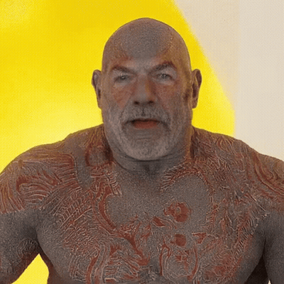A Bald Man With A Beard And A Tattoo On His Chest Looks At The Camera GIF