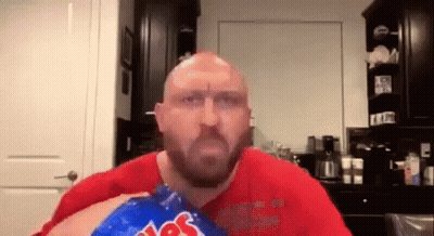A Bald Man With A Beard Is Eating A Bag Of Potato Chips In A Kitchen . GIF