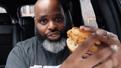 A Bald Man With A Beard Is Eating A Chicken Sandwich In A Car GIF