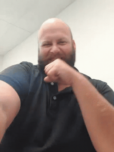 A Bald Man With A Beard Is Holding His Fist Up In The Air While Wearing A Black Shirt . GIF