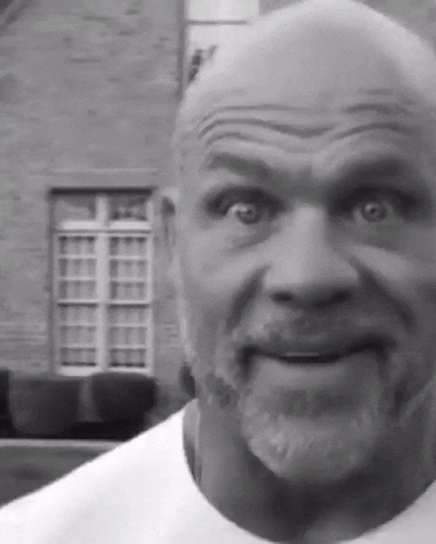 A Bald Man With A Beard Is Making A Funny Face In Front Of A Brick Building GIF