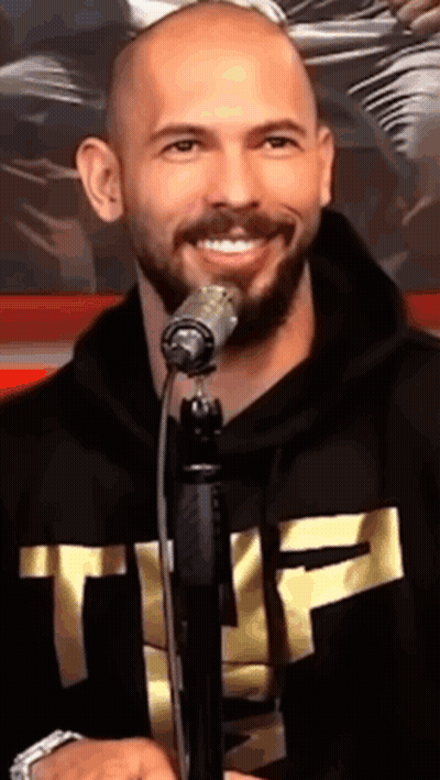 A Bald Man With A Beard Is Singing Into A Microphone While Wearing A Black Hoodie With The Letter T On It GIF