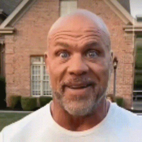A Bald Man With A Beard Is Smiling In Front Of A Brick Building . GIF