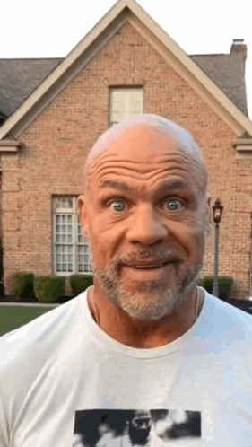 A Bald Man With A Beard Is Standing In Front Of A Brick House And Making A Funny Face GIF