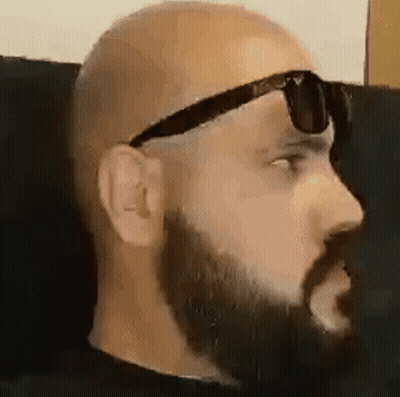A Bald Man With A Beard Is Wearing Sunglasses While Sitting On A Couch . GIF