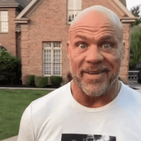A Bald Man With A Beard Wearing A White T-Shirt Is Standing In Front Of A Brick House GIF