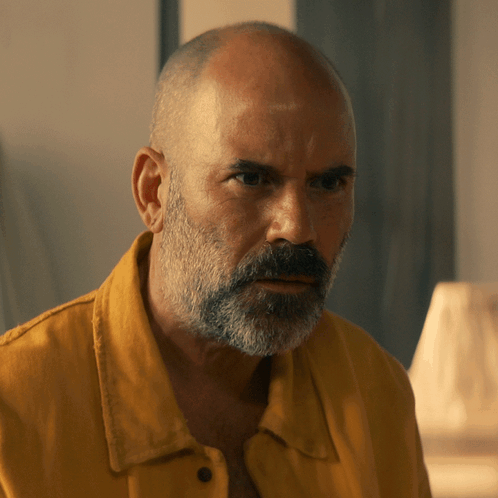 A Bald Man With A Beard Wearing A Yellow Shirt Looks At The Camera GIF