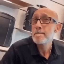 A Bald Man With A Beard Wearing Glasses And A Black Shirt Is Sitting In Front Of A Shelf Of Luggage . GIF