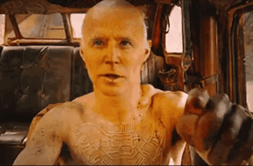 A Bald Man With A Tattoo On His Chest Is Sitting In A Car And Pointing At The Camera . GIF