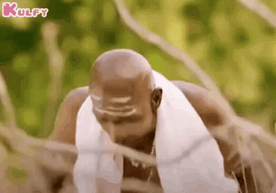 A Bald Man With A Towel Around His Neck Is Standing In A Forest . GIF