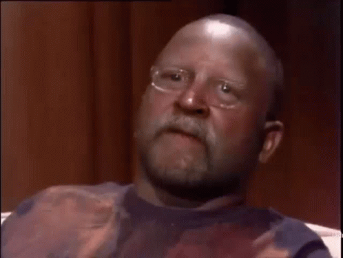 A Bald Man With Glasses And A Beard Is Sitting On A Couch Making A Funny Face . GIF