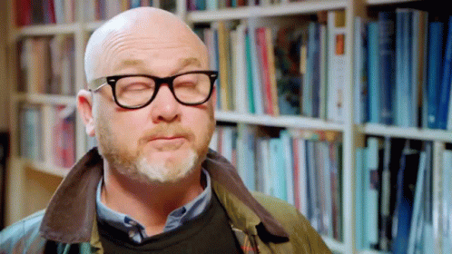 A Bald Man With Glasses And A Beard Is Standing In Front Of A Bookshelf With His Eyes Closed GIF