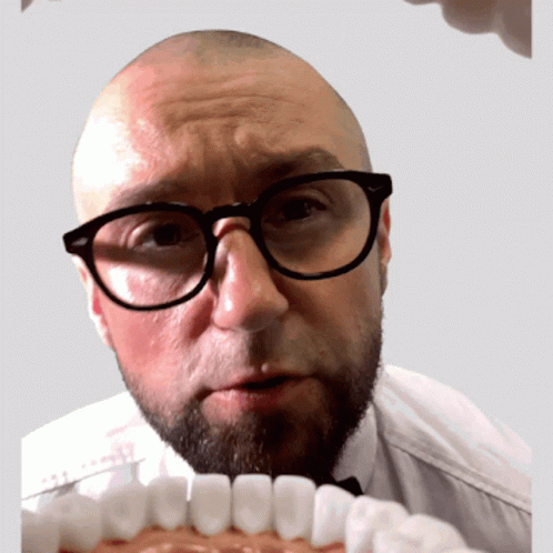 A Bald Man With Glasses And A Beard Looks At A Model Of His Teeth GIF